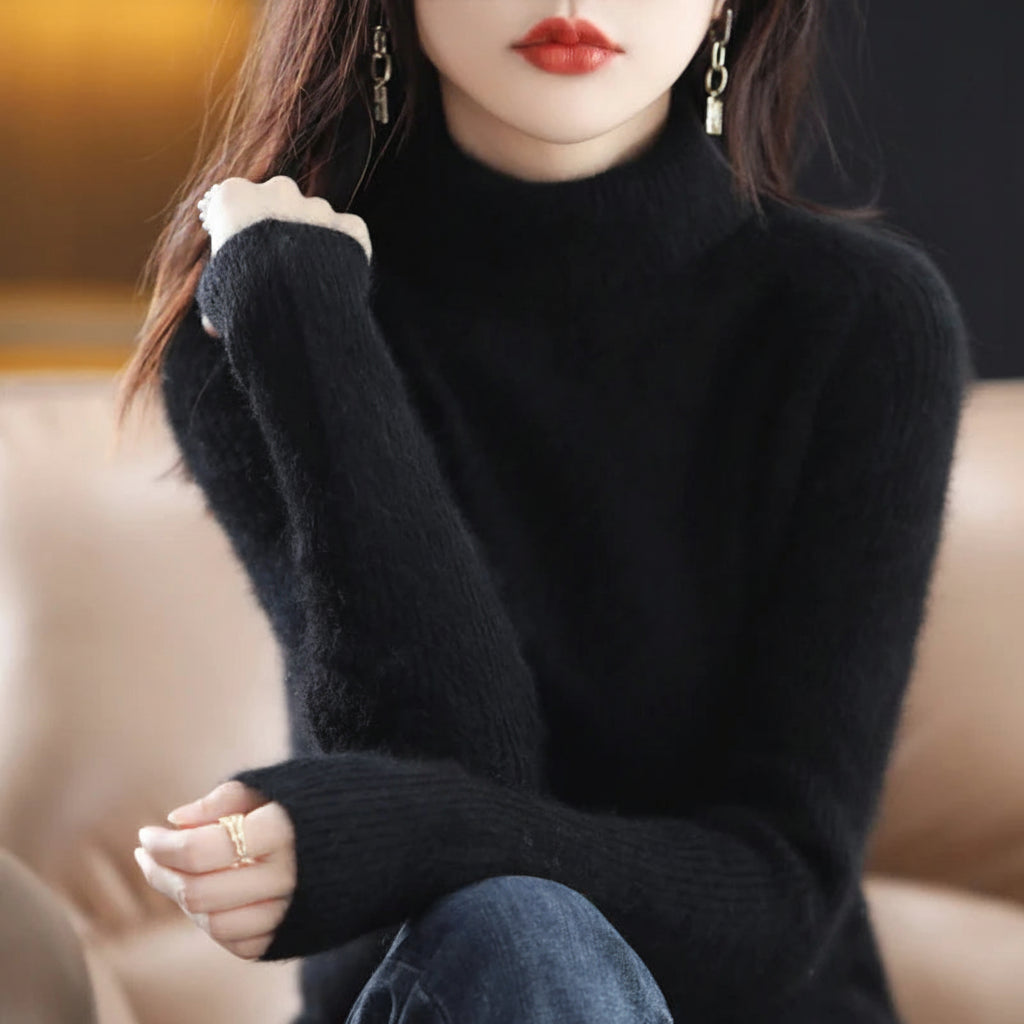 A woman wearing a black fuzzy knit mock-neck sweater, shown from the chest down with her hands crossed. She is wearing dangling earrings and a ring, seated on a beige sofa.