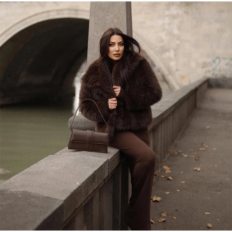 Woman in a brown fur coat sitting on a stone ledge by a body of water.