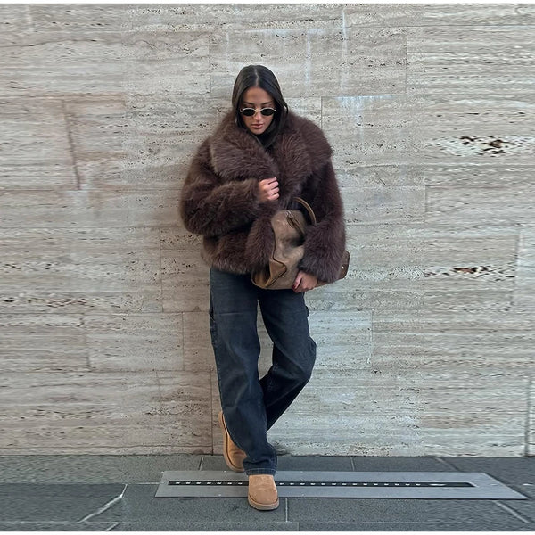 Person wearing a brown fur coat and sunglasses against a textured wall.