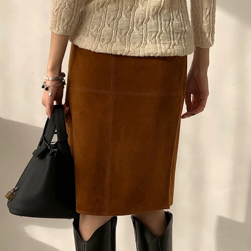 Person wearing a brown suede skirt, beige cable knit sweater, and black boots holding a black handbag.