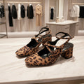 A pair of women's block-heel mules with a brown and black leopard print pattern, featuring a criss-cross strap design across the toes and thin ankle straps. The shoes are displayed on a shoe store shelf.