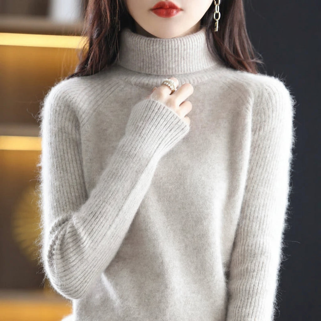 A woman wearing a light gray fuzzy turtleneck sweater, with her hand near her collarbone. The background is a dark, softly blurred interior with warm yellow lighting on the left.