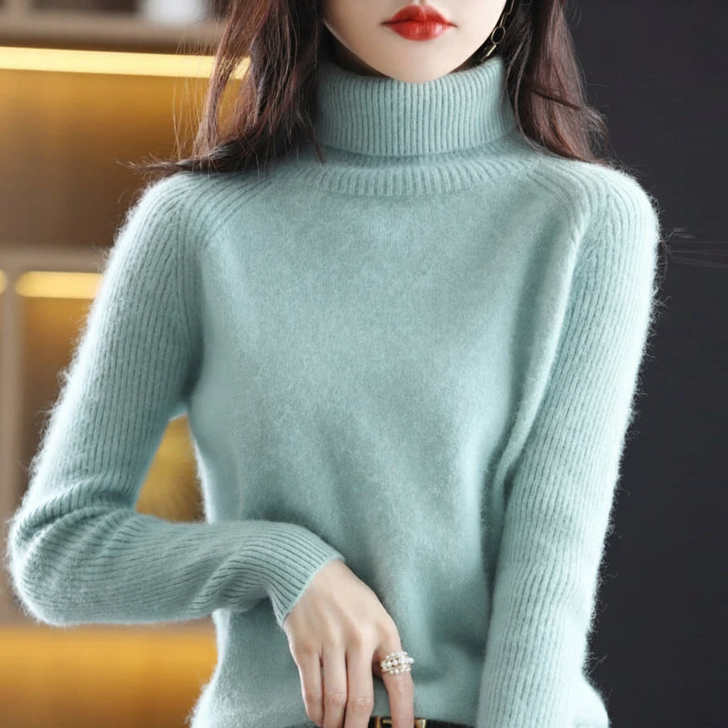 A woman wearing a light mint green turtleneck sweater, shown from the neck down. The background is a dark, softly blurred interior with warm yellow lighting.
